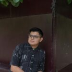 A relaxed man with glasses poses against a wall in an outdoor park in Merida, Yucatan.